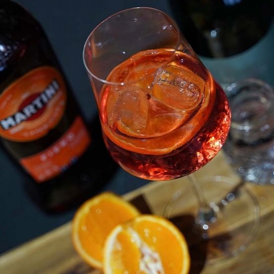 Ein Glas roter Cocktail mit Eiswürfeln und Orangenscheiben auf einer Holzoberfläche. Eine Martini-Flasche steht neben dem Glas.