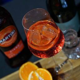 Ein Glas mit einem rosa Getränk, wahrscheinlich ein Cocktail, ist mit Eiswürfeln und Orangenscheiben serviert. Eine Flasche Martini Rosso steht links.