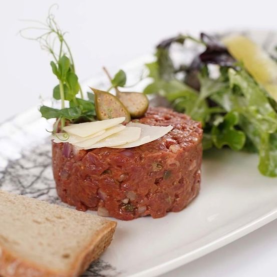 Ein Teller mit Tatar, Käse, Kräutern und einem grünen Salat, begleitet von einer Scheibe Brot.