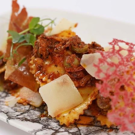 Ein Teller mit Ravioli in Tomatensoße, garniert mit Käse und grünen Kräutern, auf einem gemusterten weißen Teller.