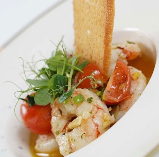 Eine weiße Schüssel mit Garnelen, Tomaten und Kräutern. Ein Stück getoastetes Brot liegt an der Seite.