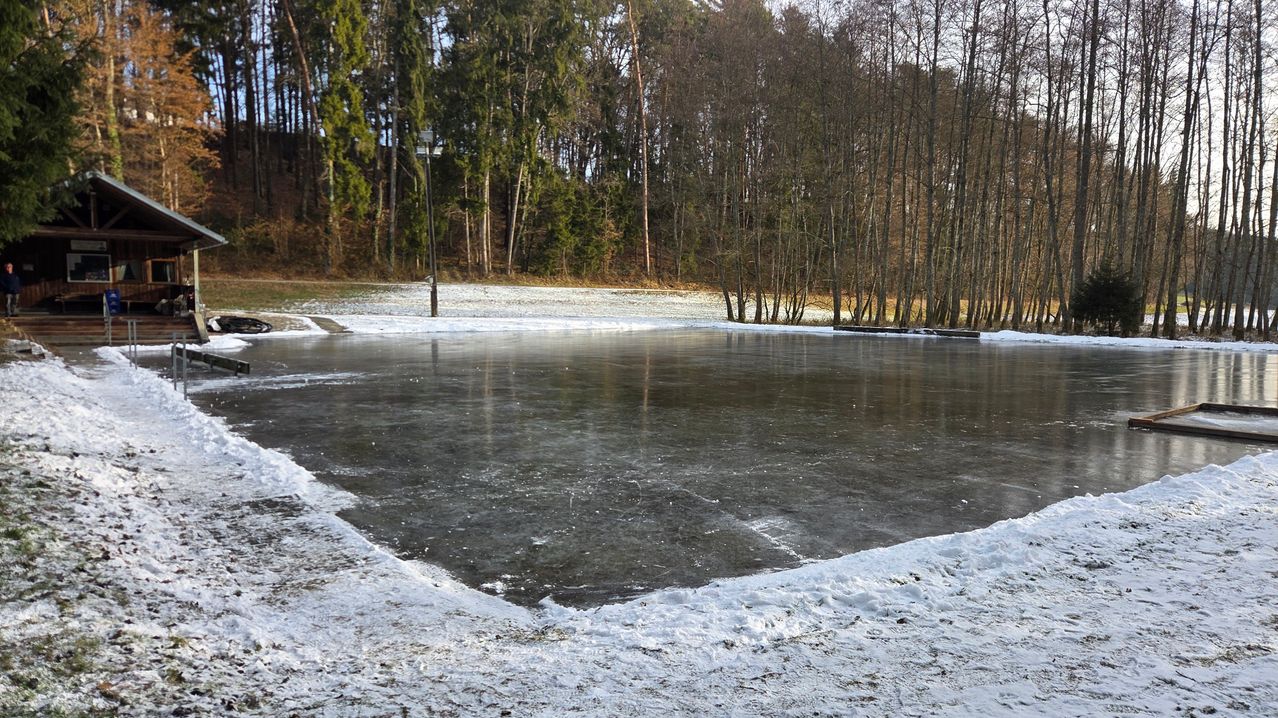 Ein zugefrorener See, umgeben von einer verschneiten Landschaft und kahlen Bäumen mit einer Straßenlaterne in der Ferne.