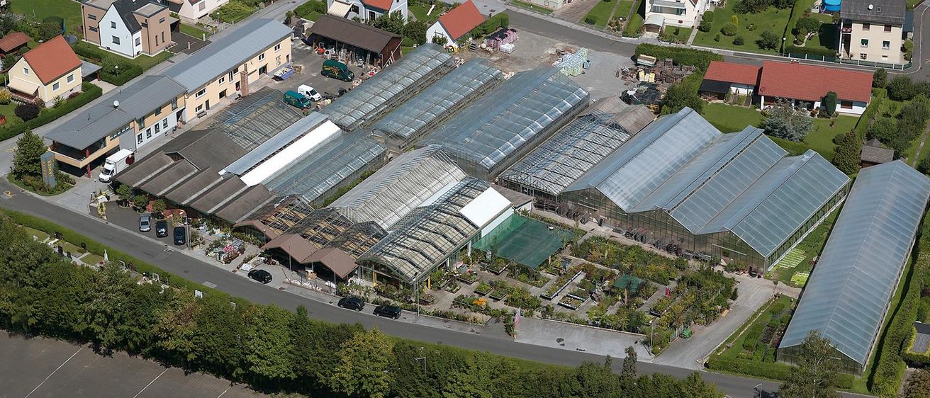 Ein Luftbild eines Gartencenters mit mehreren Gewächshäusern, geparkten Fahrzeugen und Baumaterialien. Bäume und Pflanzen sind ebenfalls sichtbar.