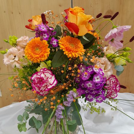 Ein farbenfroher Strauß aus verschiedenen Blumen, darunter Rosen, Nelken und Gänseblümchen, in einer durchsichtigen Glasvase auf einer weißen Tischdecke angeordnet.