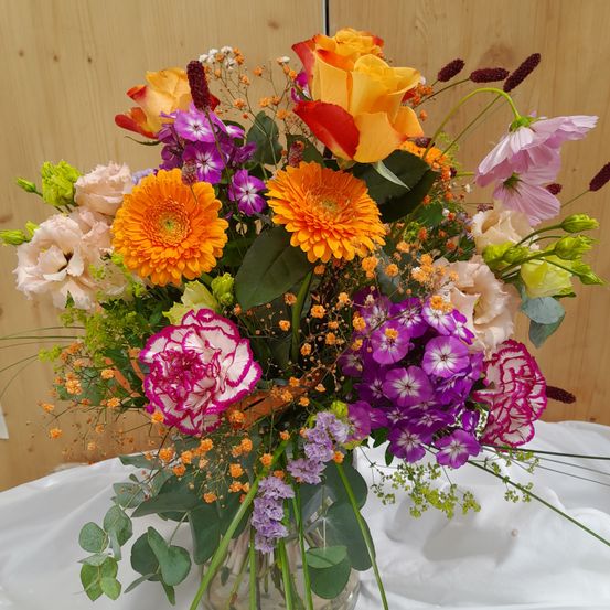 Ein farbenfroher Strauß aus verschiedenen Blumen, darunter Rosen, Nelken und Gänseblümchen, in einer durchsichtigen Glasvase auf einer weißen Tischdecke angeordnet.