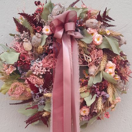 Ein Kranz aus getrockneten Blumen und Grün hängt an einer weißen Wand. Die Blumen sind rosa, lila und gelb, und die Blätter sind grün. Der Kranz hat eine rosa Schleife, die daran gebunden ist.