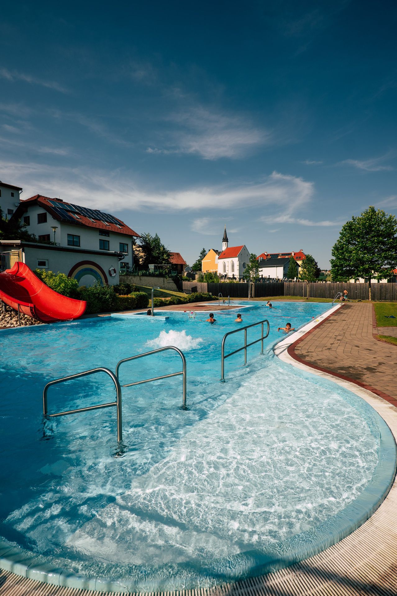 Ein Freibad mit schwimmenden Personen, umgeben von einem Zaun. Ein roter Wasserrutsche ist in der Nähe. Ein Haus mit Solarmodulen und einem Regenbogendesign ist im Hintergrund.