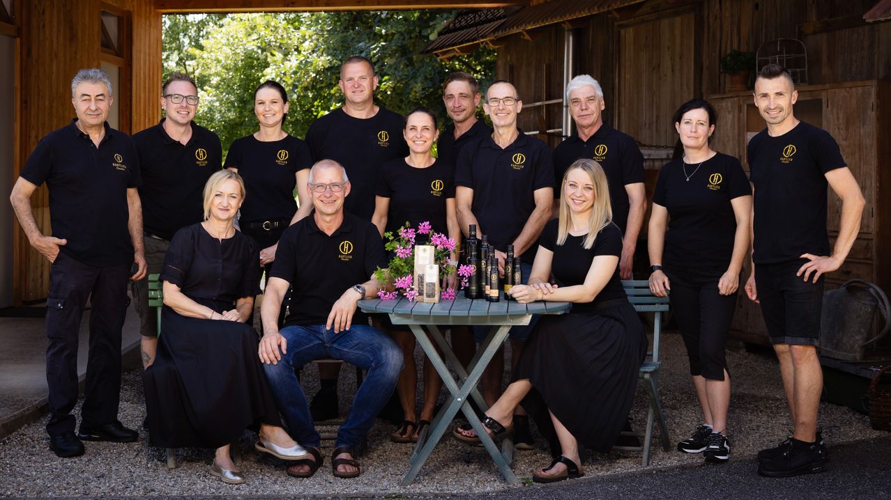 A group of people in matching black shirts with yellow logos are posing for a photo in front of a rustic building. They are sitting and standing around a table with bottles and flowers.