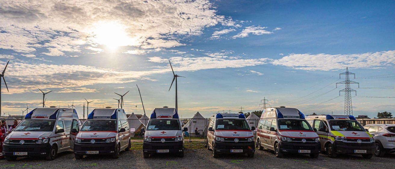 Fünf Krankenwagen sind auf einem Kiesplatz geparkt. Sie sind weiß und rot. Im Hintergrund befinden sich einige Windräder und Stromleitungen.