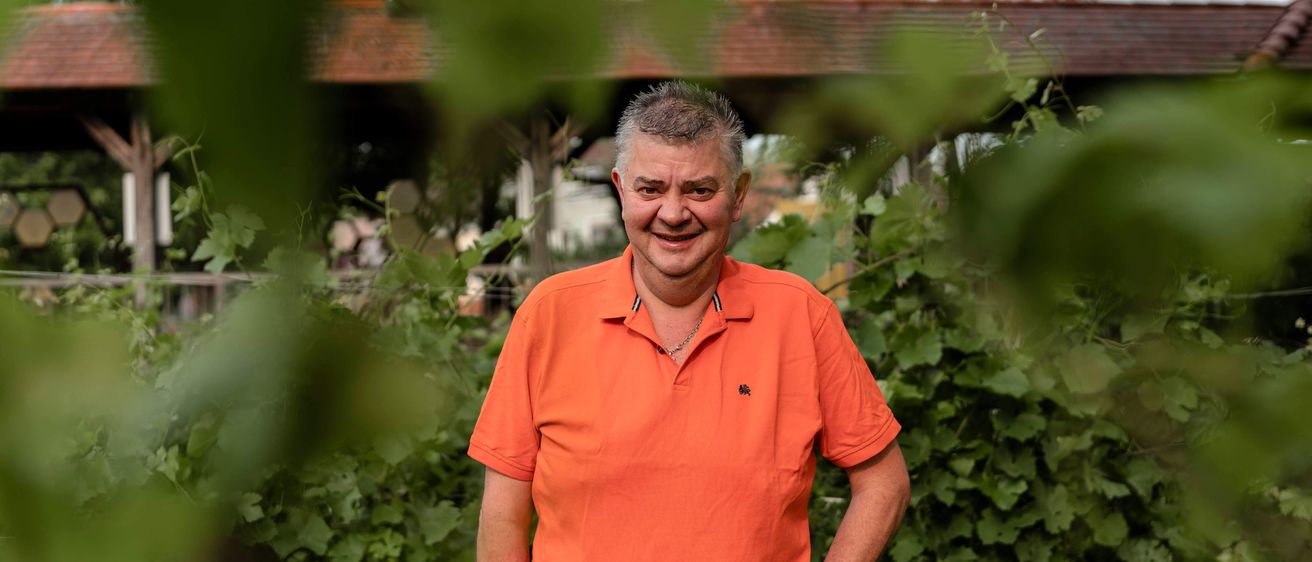 Ein Mann in einem orangefarbenen Polohemd steht in einem Garten, lächelt und hat grünes Laub im Hintergrund.