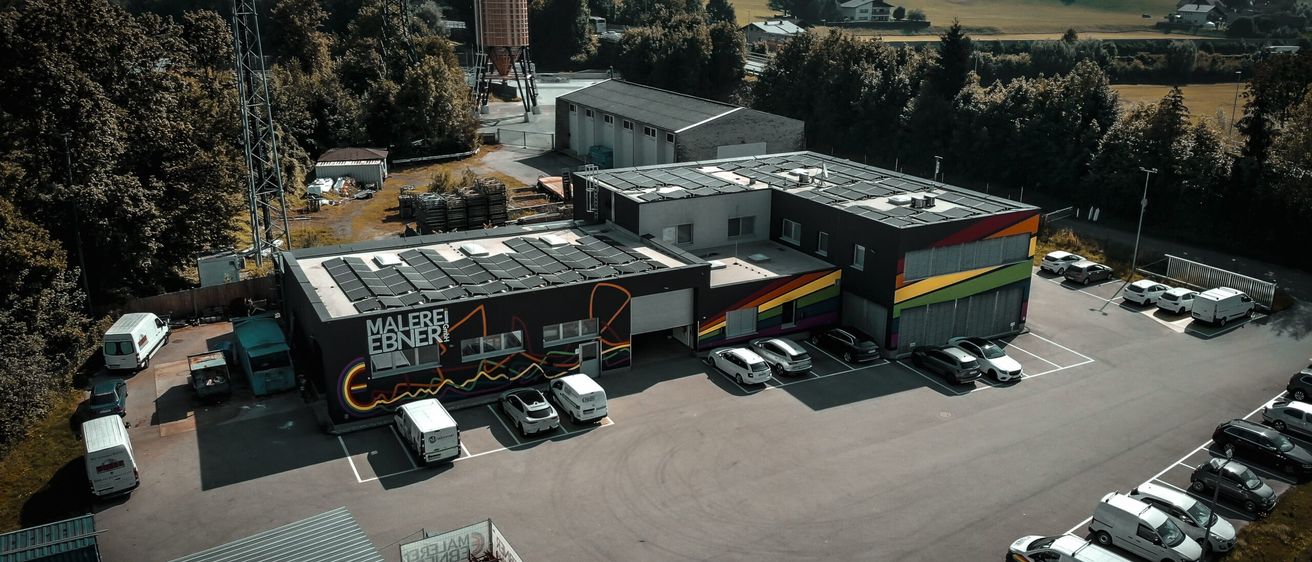 Eine Luftaufnahme eines großen Gebäudes mit Solarpanelen, bunten Wandgemälden und einem Parkplatz mit mehreren geparkten Autos.
