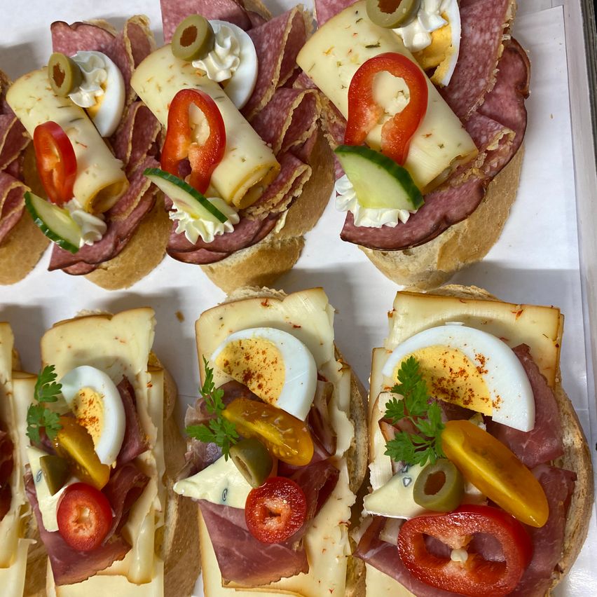 Nahaufnahme mehrerer Sandwichhälften mit verschiedenen Belägen wie Eiern, Käse, Oliven und Tomaten, die auf einer weißen Oberfläche angeordnet sind.