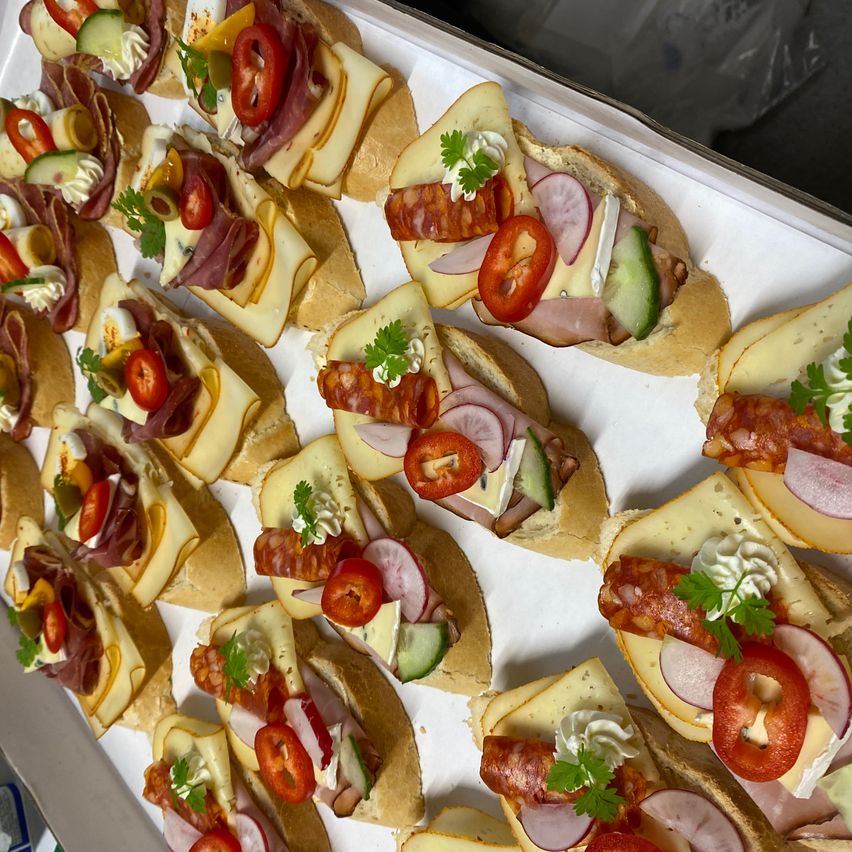 Ein Tablett gefüllt mit verschiedenen Sandwiches, jedes belegt mit Käsescheiben, Tomaten, Gurken und Fleisch. Mit Petersilie garniert sind die Sandwiches ordentlich und appetitlich angeordnet.