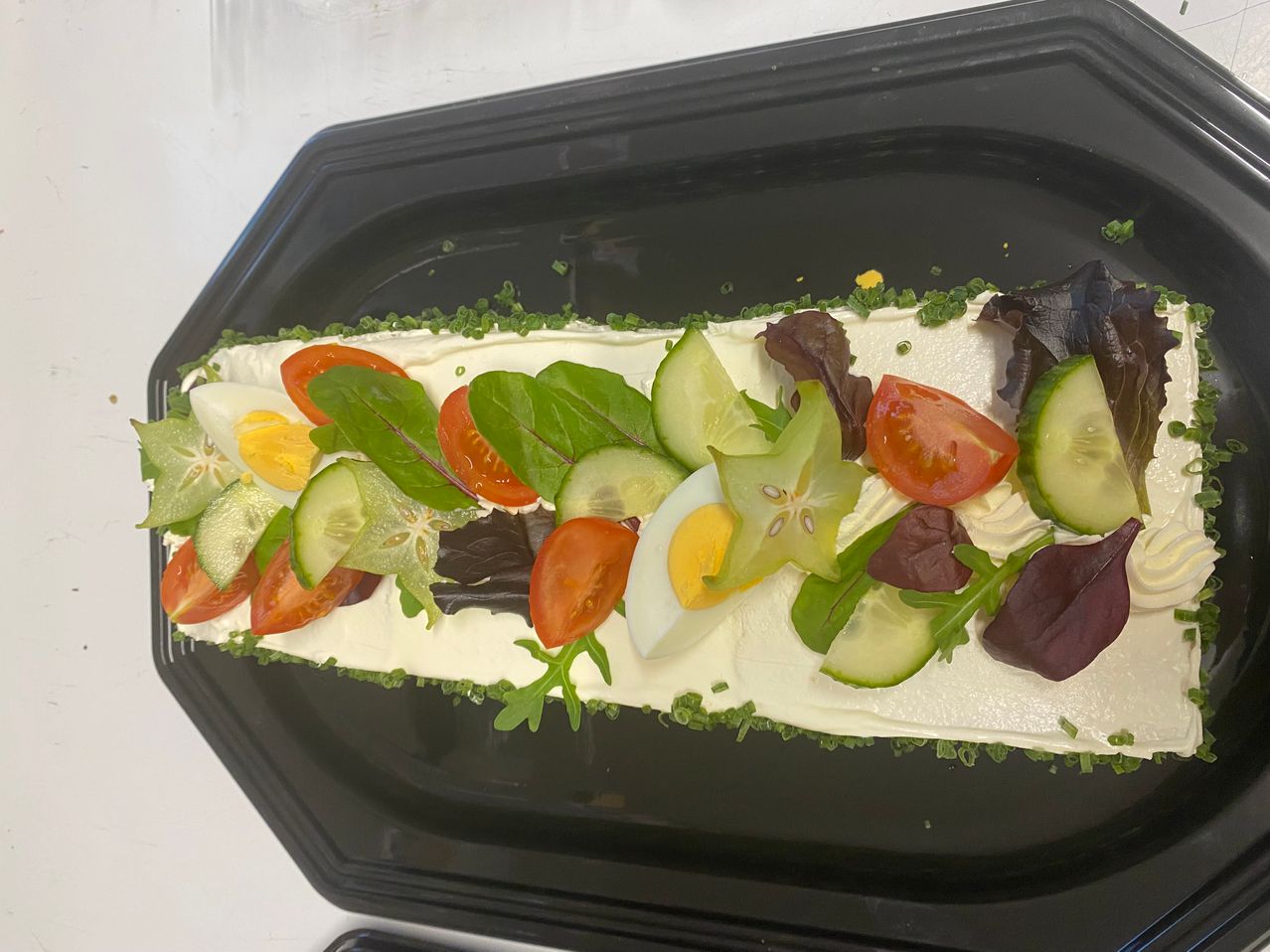 Eine Gemüseplatte auf einer weißen Sahnebasis, verziert mit Scheiben von Gurke, Tomate, Ei und verschiedenen Blattgemüsen. Die Platte ist auf einem schwarzen rechteckigen Tablett arrangiert.