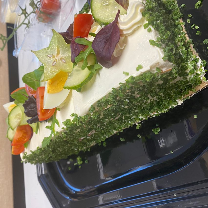 Ein Sandwich mit weißer Frischkäsecreme, belegt mit Gurke, Tomate, Sternfrucht und grünen Kräutern auf einem schwarzen Teller.