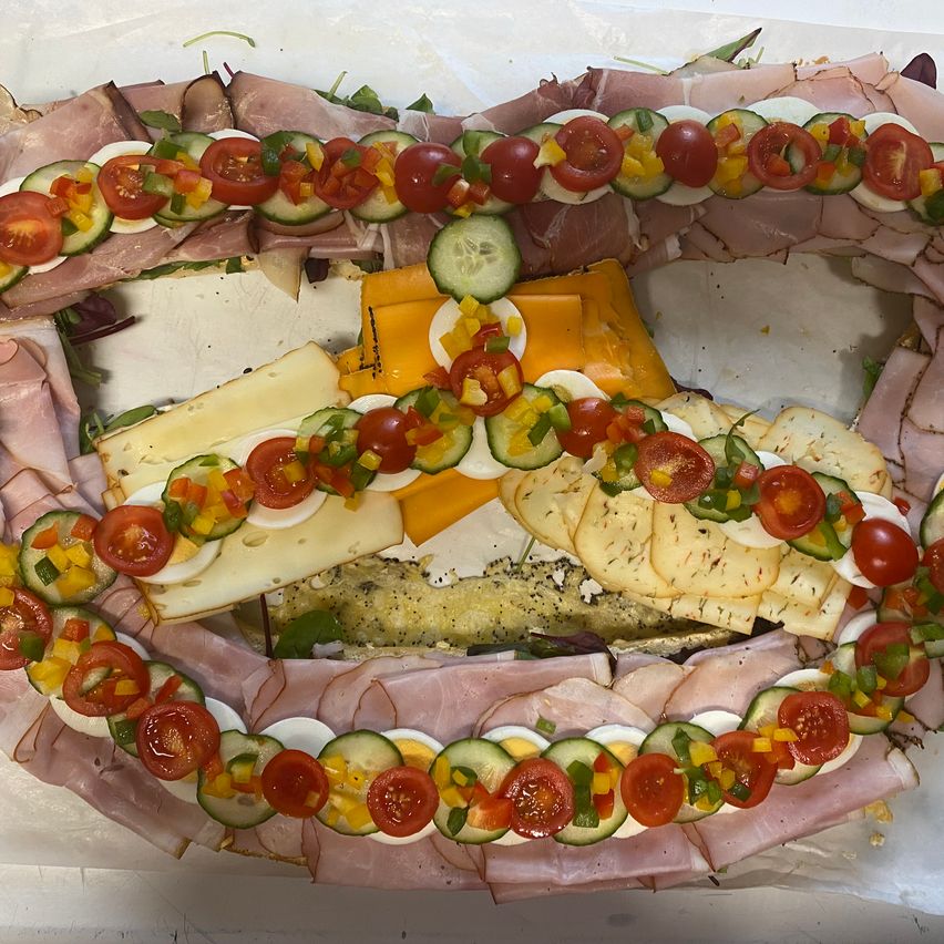 Eine kreisförmige Platte zeigt eine Vielzahl von geschnittenem Fleisch, Käse und Gemüse, darunter Tomaten, Gurken und Eier, in einer dekorativen Anordnung.
