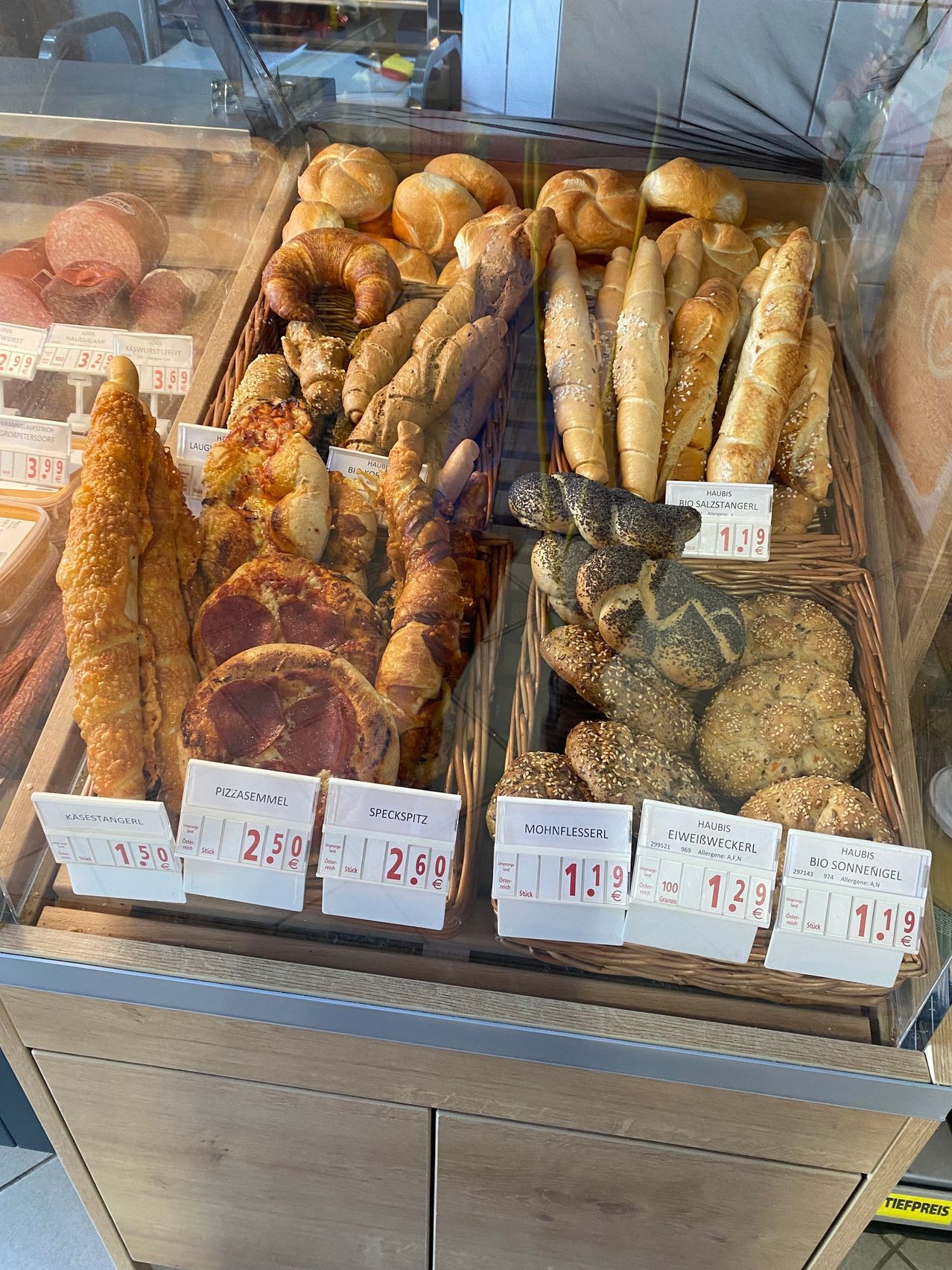 A display case filled with various types of bread and pastries, including croissants, pretzels, and bread rolls, all neatly arranged in baskets with price tags.