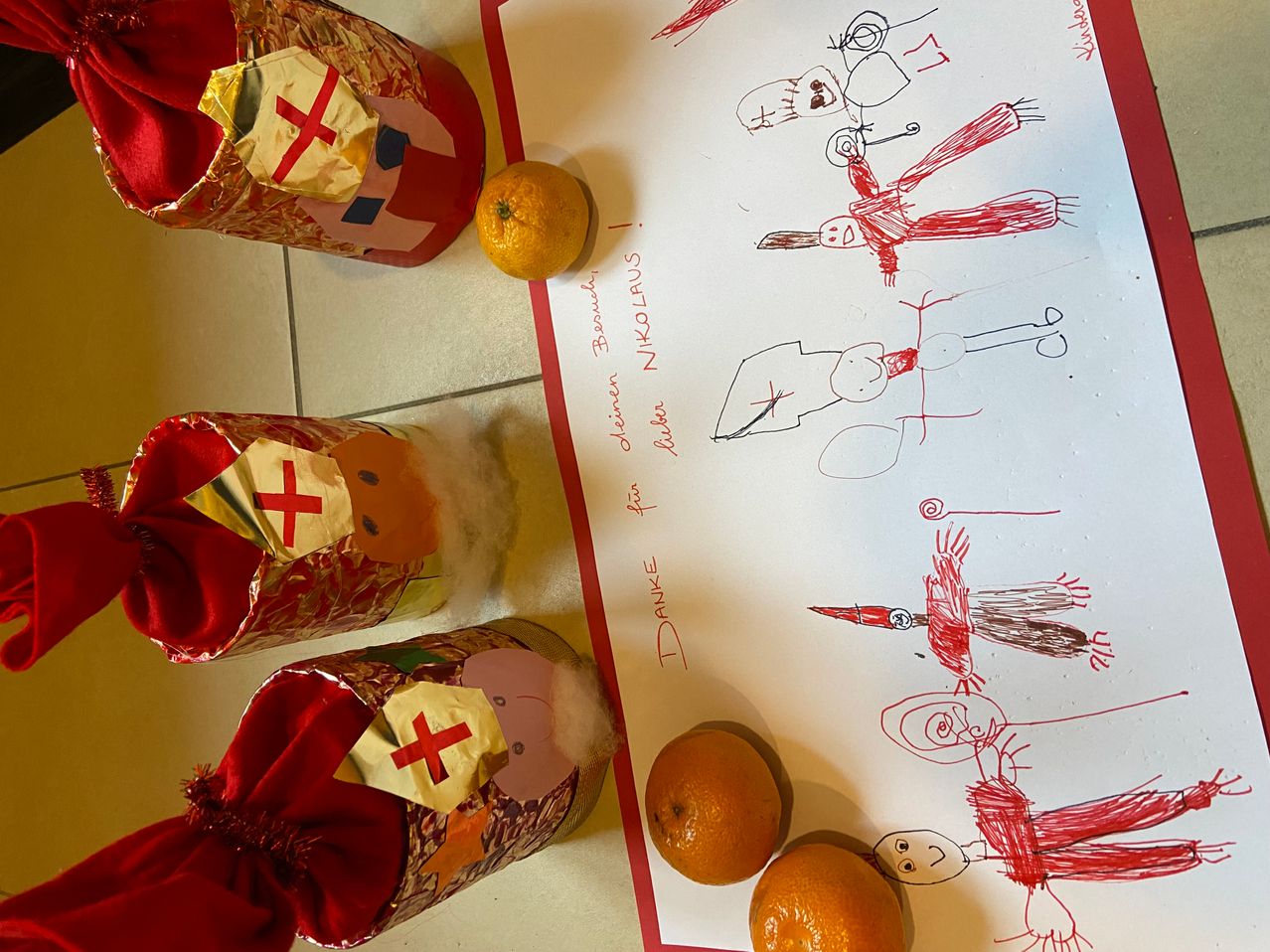 Eine Zeichnung auf weißem Papier mit roten Linien zeigt verschiedene Figuren. Drei rote Schuhkartons mit goldenen und roten Dekorationen befinden sich neben dem Papier. Drei Orangen liegen auf dem Papier.