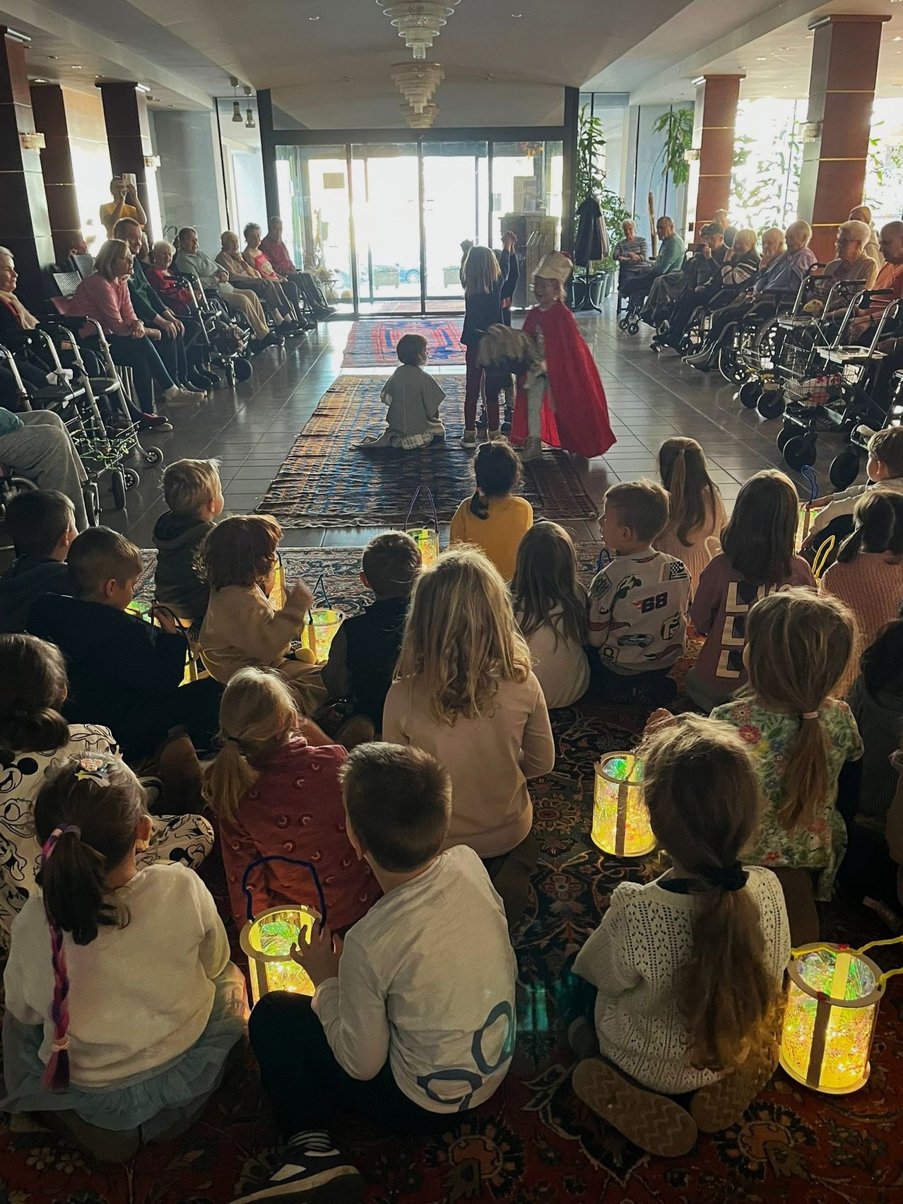 Eine Gruppe von Kindern und Erwachsenen sitzt auf dem Boden und beobachtet eine Person in einem roten Umhang und Hut auf einem Teppich. Einige halten Laternen und andere sind in Rollstühlen. Einige Leute sitzen auf Stühlen auf der linken Seite.