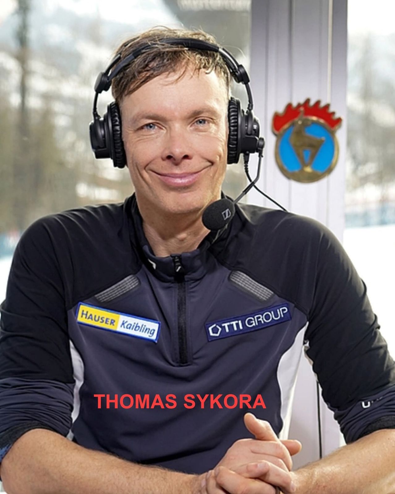 Ein lächelnder Mann mit Headset und einer Jacke mit Logos sitzt vor einem Mikrofon. Sein Name ist Thomas Sykora.