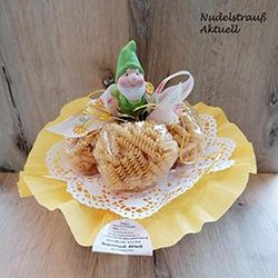 Ein Teller mit einer Zwergenfigur auf einem Stapel Pasta, geschmückt mit einem gelben Spitzendeckchen und einem kleinen Etikett vorne. Der Teller steht auf einer Holzoberfläche.