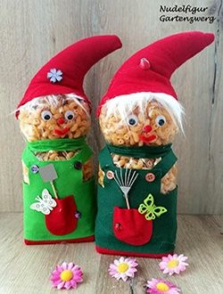 Zwei Zwerge aus Popcorn und grünem Stoff, mit roten Mützen, grünen Westen und Werkzeugen, auf einer Holzoberfläche sitzend, umgeben von Blumen.