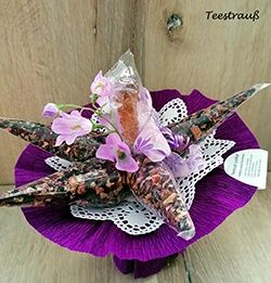 Eine sternförmige Anordnung mit getrockneten Blumen und Kräutern in Plastiktüten auf einem violetten Spitzendeckchen auf einer Holzoberfläche.