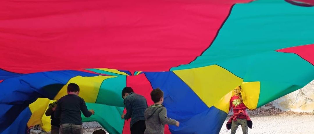 Fünf Jungen stehen unter einem farbenfrohen großen Luftballon. Ein Junge in einem roten Kostüm hält den Ballon, während die anderen versuchen, hineinzukommen.