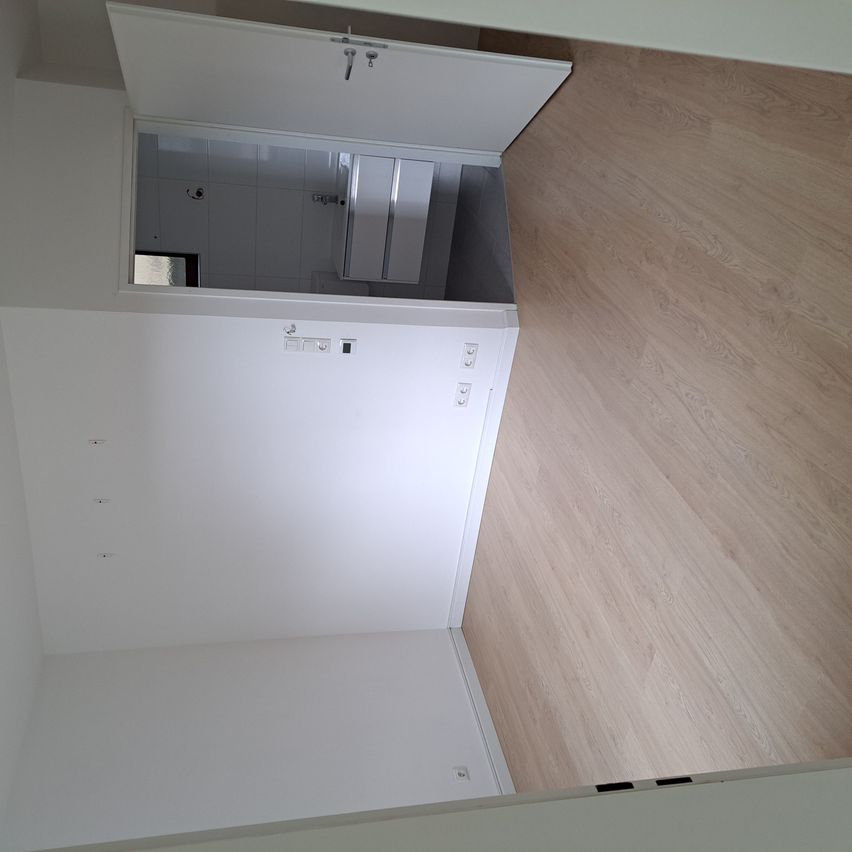 Eine Draufsicht eines leeren Raums mit weißen Wänden und Holzböden. Eine Tür ist offen und führt in einen kleineren Raum mit einem Spiegel. Steckdosen und Schalter sind an den Wänden sichtbar.