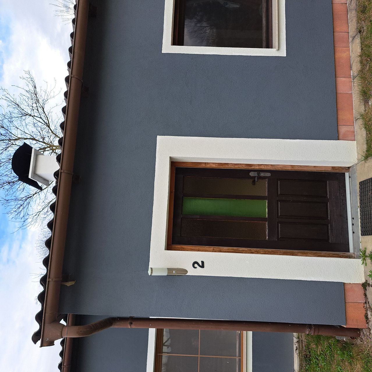 Eine Vogelperspektive eines grauen Hauses mit einer braunen Tür und zwei Fenstern. Die Nummer 2 ist über der Tür angebracht.