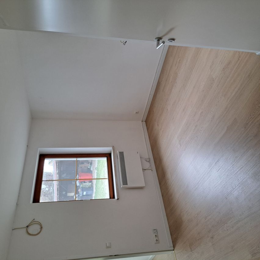 Ein leerer Raum mit weißen Wänden und Holzboden. Ein kleines Fenster ist an der linken Wand und eine Klimaanlage ist an der Wand über dem Fenster montiert. Eine Steckdose befindet sich an der Wand unter dem Fenster.