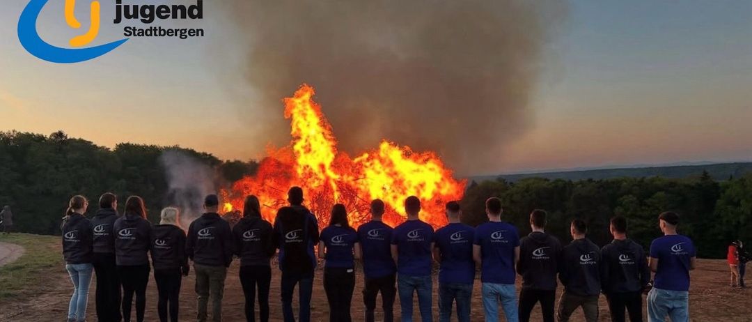Eine Gruppe von Menschen in blauen Shirts steht vor einem großen Lagerfeuer bei Dämmerung. Im Hintergrund sind Bäume und ein klarer Himmel zu sehen.