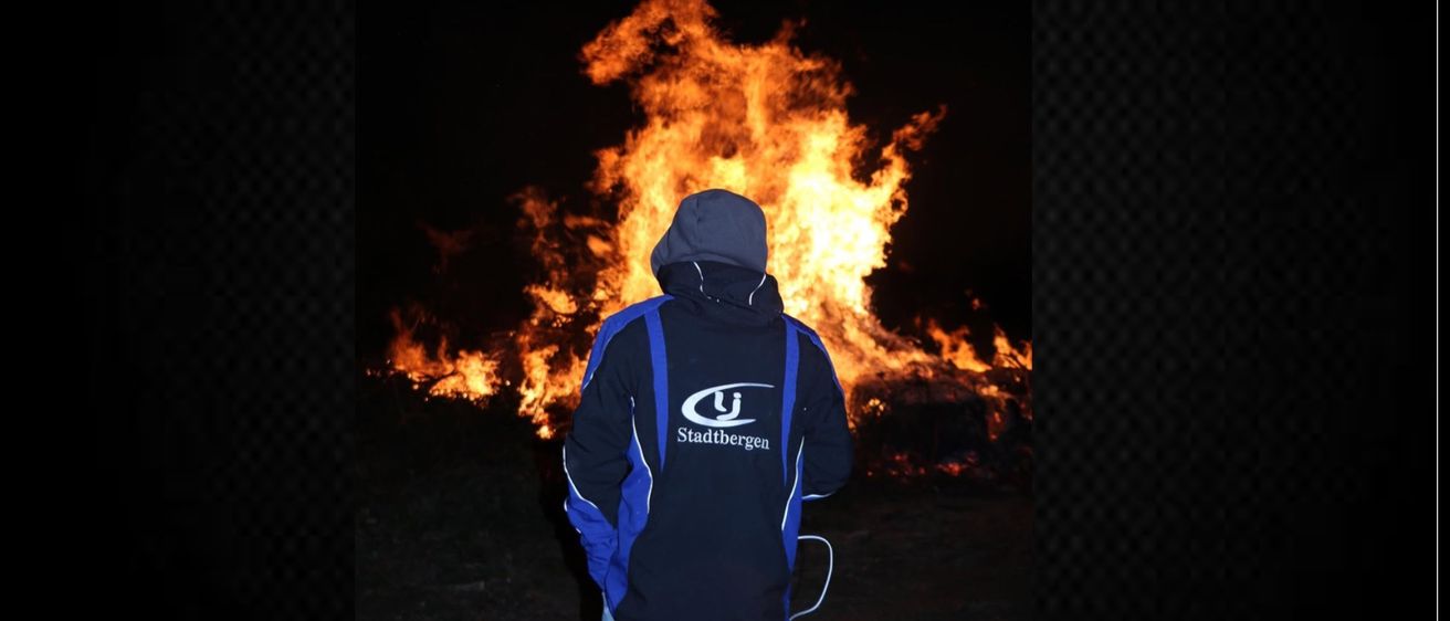 Eine Person in einer blau-schwarzen Jacke steht bei Nacht vor einem großen Lagerfeuer.
