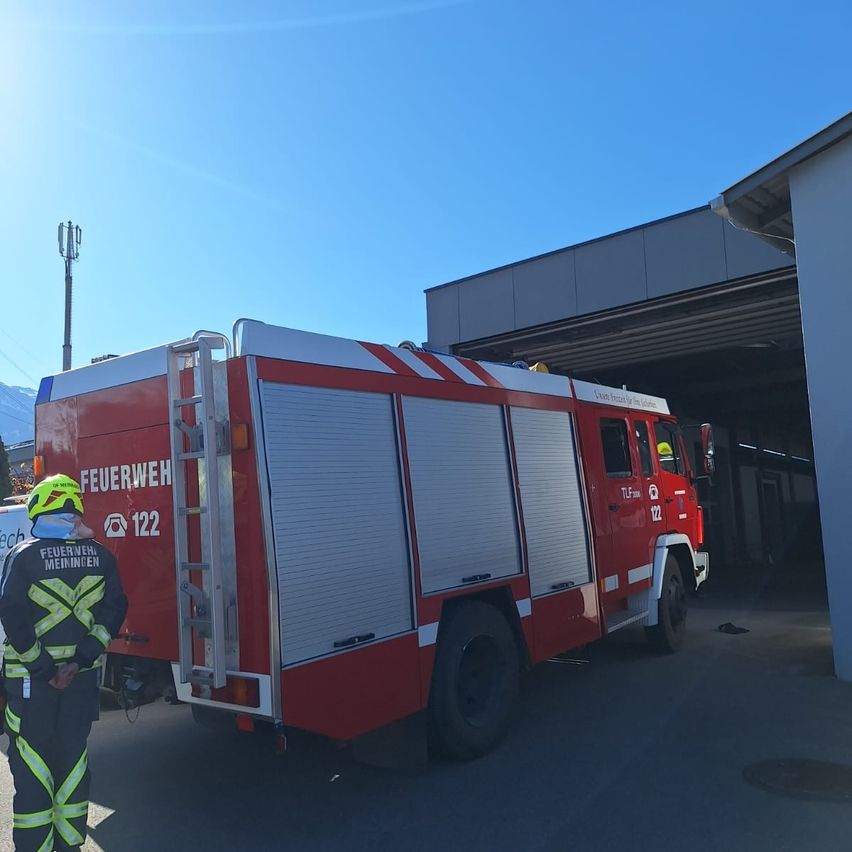 Ein Feuerwehrmann steht neben einem roten Feuerwehrauto, Nummer 122, das vor einem Gebäude geparkt ist. Der Himmel ist blau mit einigen Wolken.