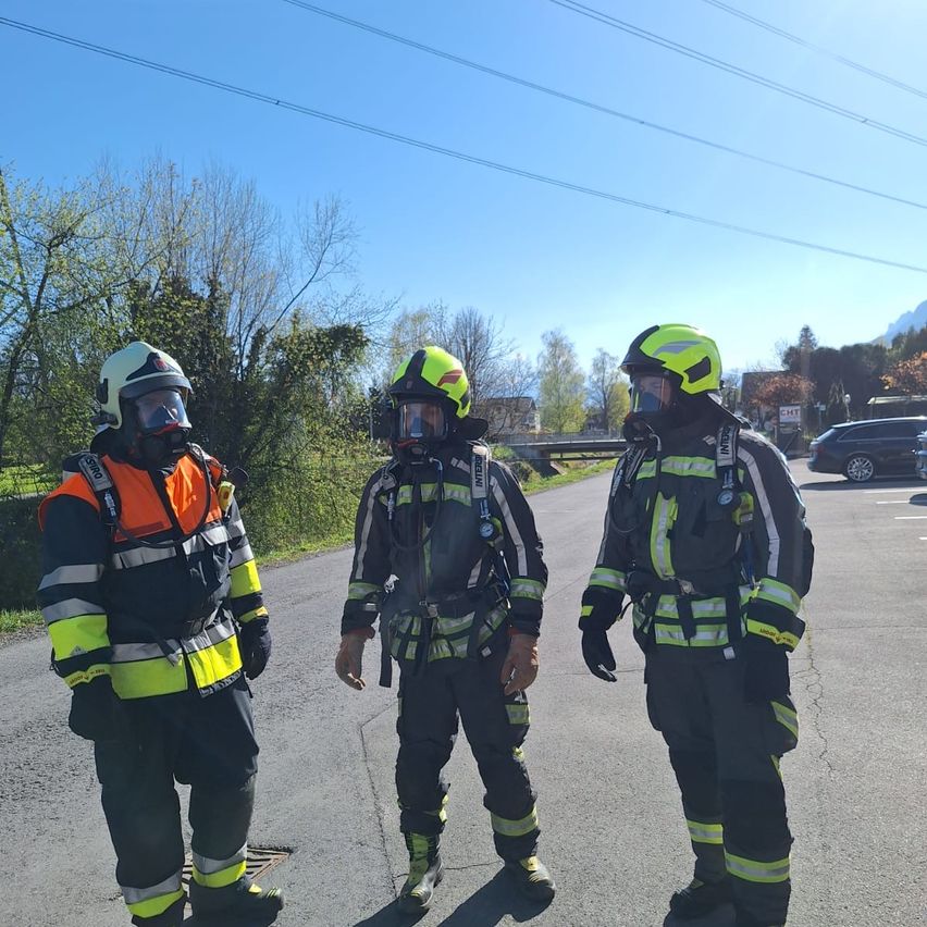Drei Feuerwehrleute in voller Ausrüstung stehen auf einer Straße mit grünen und gelben Markierungen, unter einem blauen Himmel mit Wolken. Sie sind mit Helmen und Masken ausgestattet.