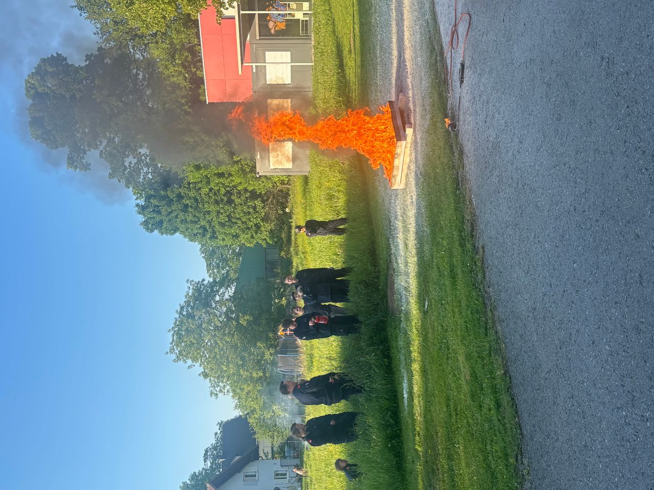 Eine Gruppe von Menschen in Schwarz beobachtet ein Feuer in der Nähe eines Gebäudes mit rotem Dach. Bäume und Gras umgeben die Szene, und eine Straße ist rechts sichtbar.