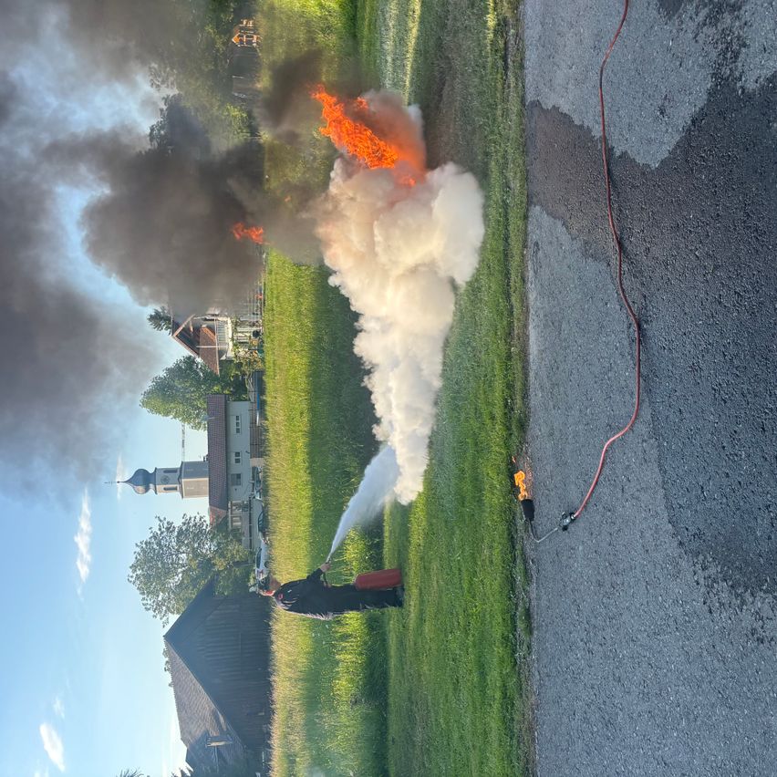 Ein Feuerwehrmann kämpft mit einem Feuerlöscher gegen ein Feuer in einem Grasfeld. Rauch und Flammen steigen aus dem brennenden Bereich auf. Eine Feuerwehrschläuche liegt in der Nähe.