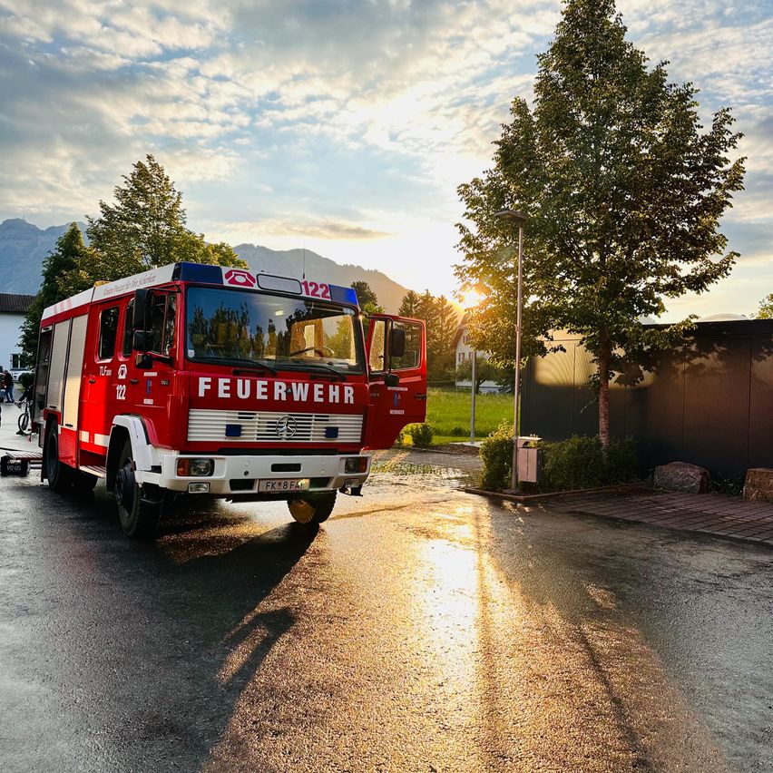 Ein roter Feuerwehrwagen steht auf einer nassen Straße, mit geöffneter Tür und Bergen im Hintergrund. Bäume und Gebäude umgeben das Gebiet.