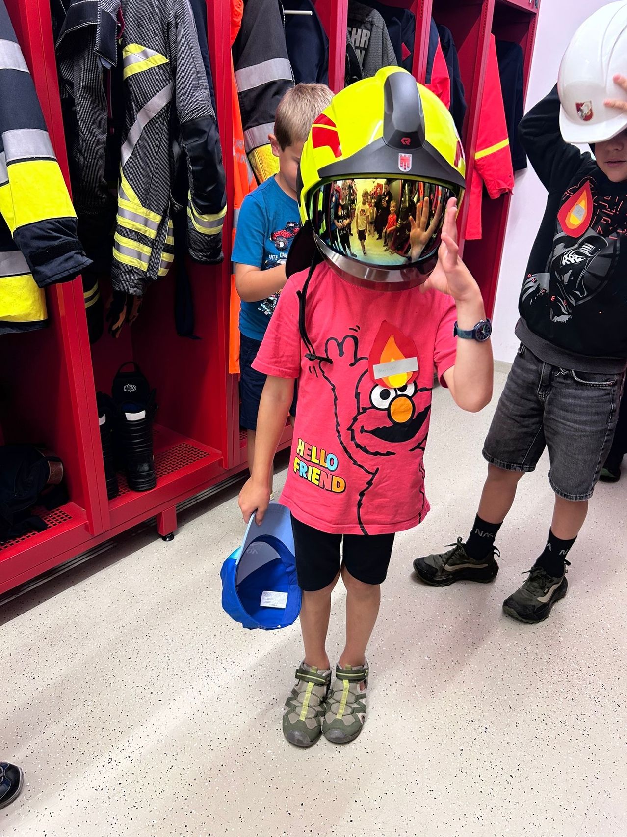 Ein junger Junge mit einem Feuerwehrhelm steht in einem Umkleideraum, hält einen blauen Hut in der Hand, mit zwei anderen Kindern in der Nähe. Hinter ihm befinden sich rote Spinde mit Feuerwehrjacken, die hängen.