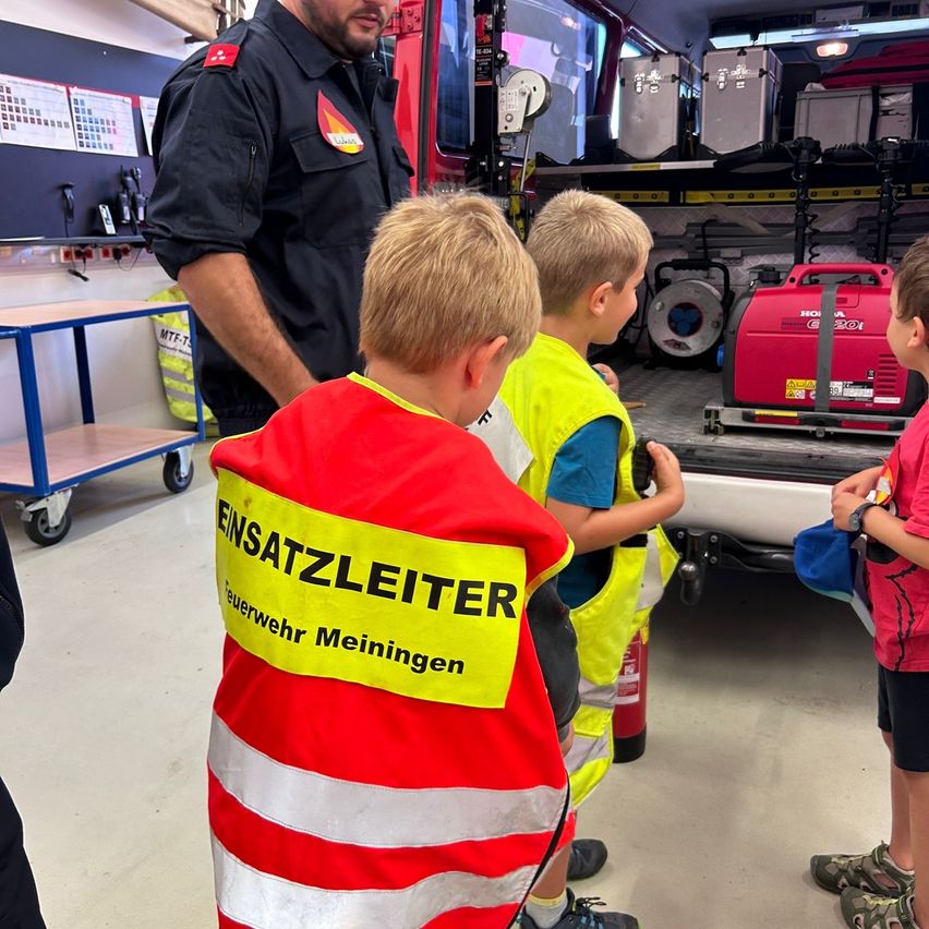 Ein Feuerwehrmann in Uniform steht neben einem Feuerwehrauto und weist drei Kinder an, von denen eines eine Warnweste trägt.