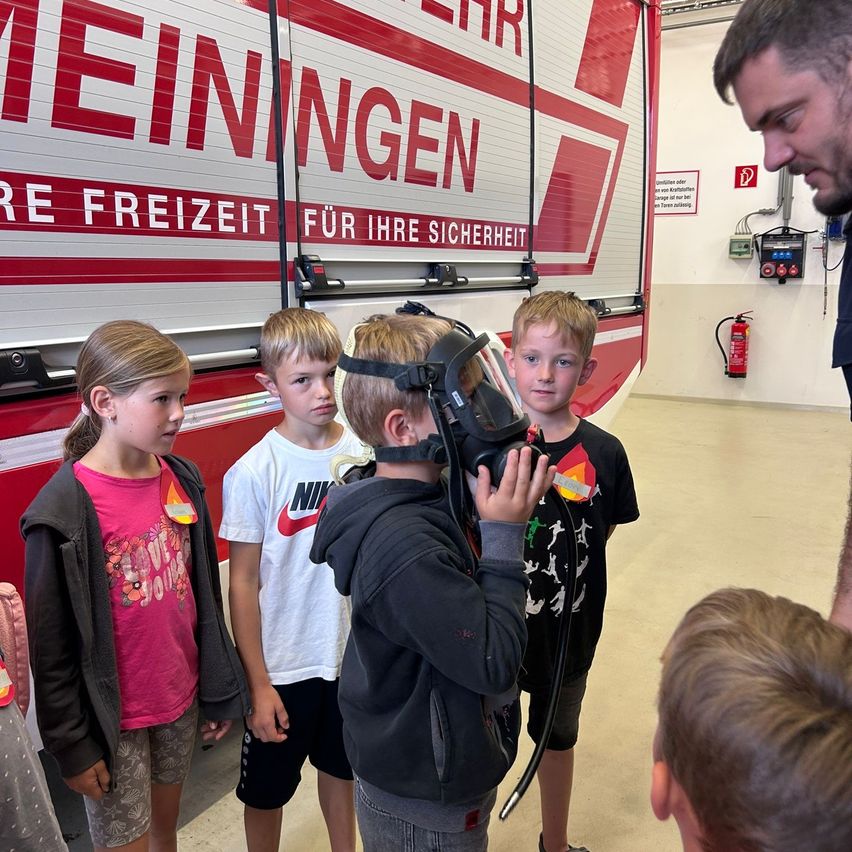 Fünf Kinder befinden sich in einer Feuerwache. Ein Junge trägt eine Gasmaske. Ein Mann in Uniform steht neben ihnen und gibt Anweisungen. Im Hintergrund ist der Feuerwehrwagen prominent zu sehen.