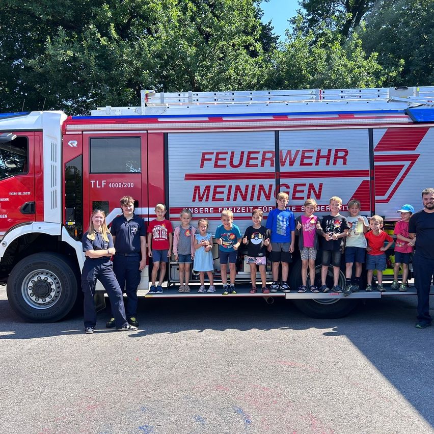 Eine Gruppe von Kindern und Erwachsenen steht auf der Rückseite eines roten Feuerwehrwagens mit dem Schriftzug 'Feuerwehr Meiningen'. Der Wagen ist auf einer Straße geparkt, mit Bäumen im Hintergrund.
