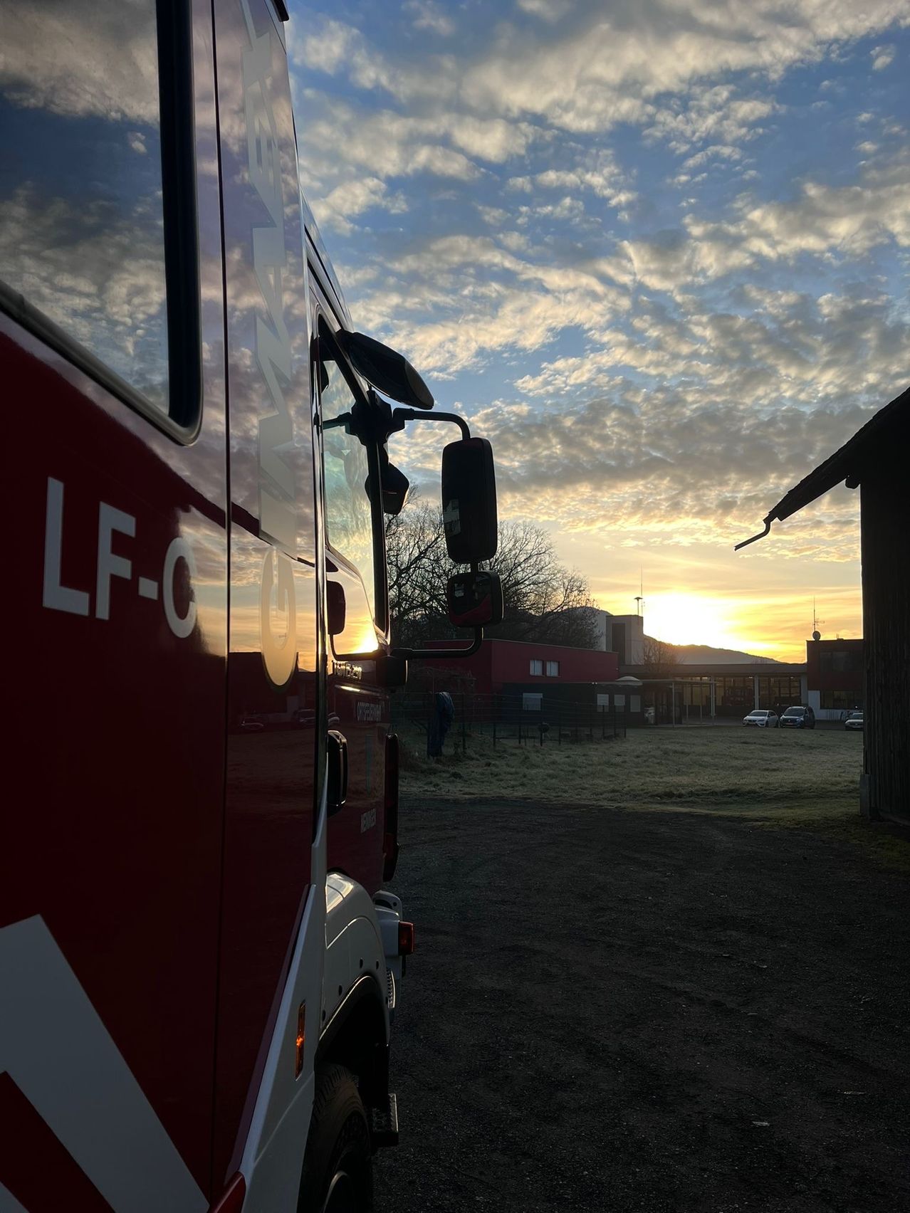 Ein rotes Fahrzeug, wahrscheinlich ein Feuerwehrauto, steht auf einem offenen Gelände. Dahinter ein Gebäude mit rotem Dach. Der Himmel ist bewölkt mit untergehender Sonne.