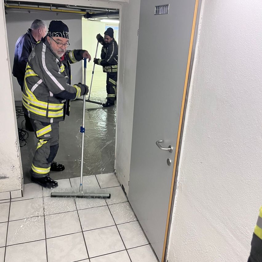 Drei Feuerwehrleute in Uniform reinigen den Boden eines Gebäudes mit Mopps und Besen. Einer hält einen Mopp und reinigt den Boden. Ein anderer steht mit einem Besen. Der Raum hat weiße Fliesenböden und Wände.
