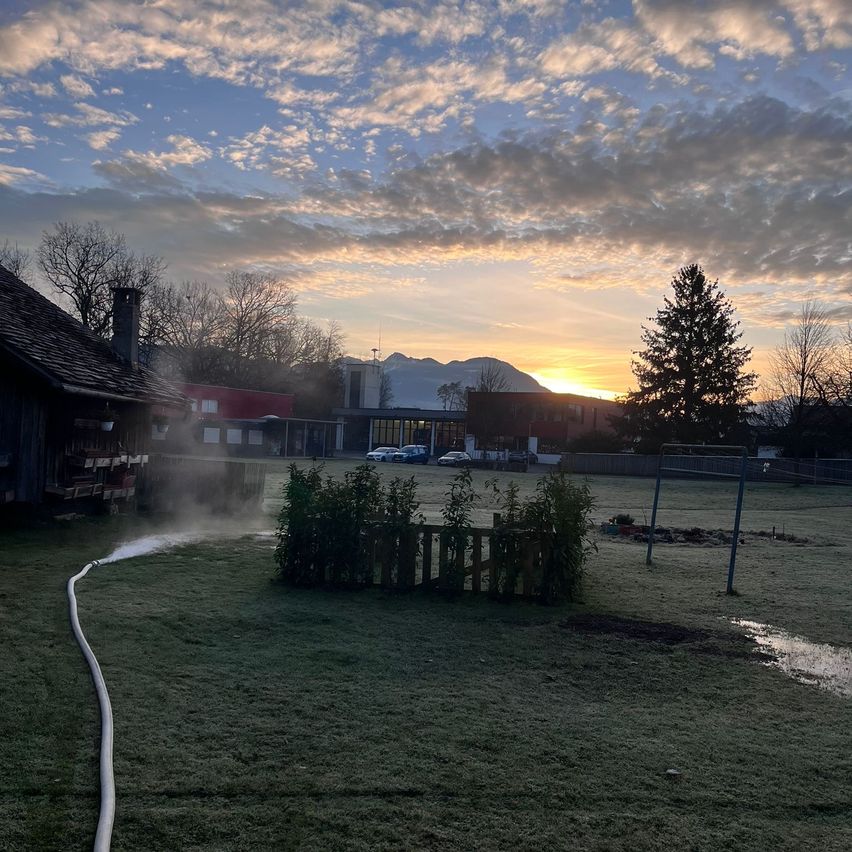 Bei Sonnenuntergang ein Feld mit Zaun und Gebäude. Bäume und Berge im Hintergrund. Autos vor dem Gebäude geparkt. Rauch kommt aus einem Schornstein.