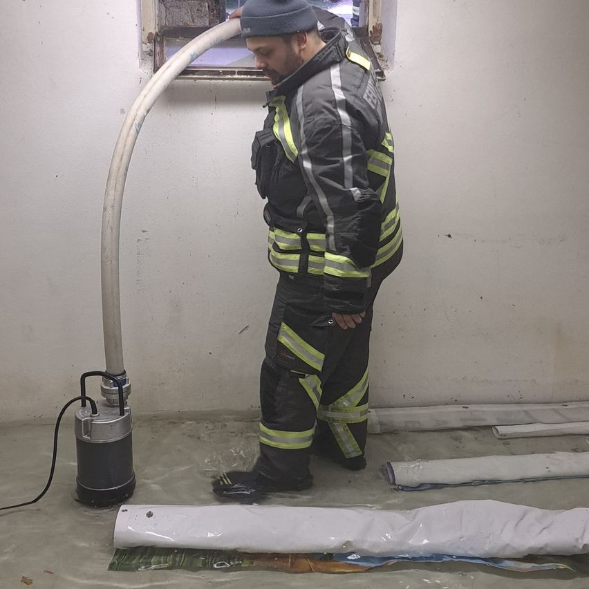 Ein Feuerwehrmann in Uniform steht in einem überfluteten Keller und benutzt eine Pumpe. Eine große Rohrleitung ist an die Pumpe angeschlossen. Die Wände sind nass und der Boden ist mit Wasser bedeckt.