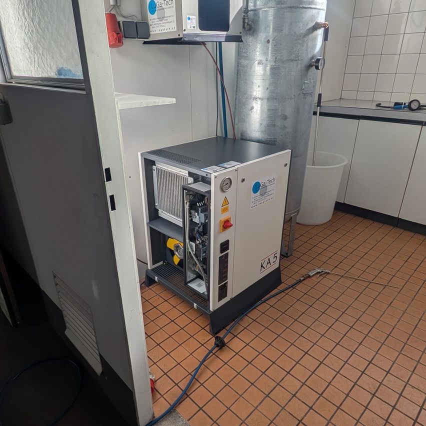 Ein industrielles Umfeld mit einer KA 5 Maschine, einem großen zylindrischen Tank und einem Schrank mit einer Mikrowelle oben. Ein blauer Schlauch ist an der Maschine angeschlossen.