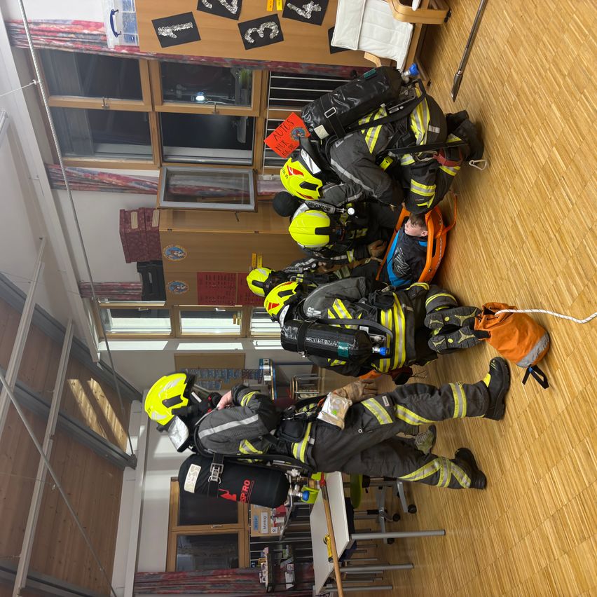 Eine Gruppe von Feuerwehrleuten in Schutzausrüstung übt eine Evakuierung auf einem Holzboden. Eine Person liegt auf einer Trage, andere stehen oder knien um sie herum. Der Raum hat Holzschränke, Glasfenster und einen weißen Tisch.