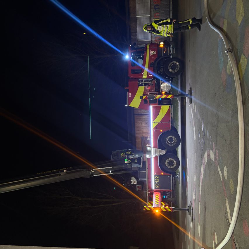 Ein Feuerwehrauto ist nachts am Straßenrand geparkt. Die Lichter des Autos sind eingeschaltet, und der Fahrer steht auf der Leiter. Der Körper des Autos ist rot und gelb lackiert.