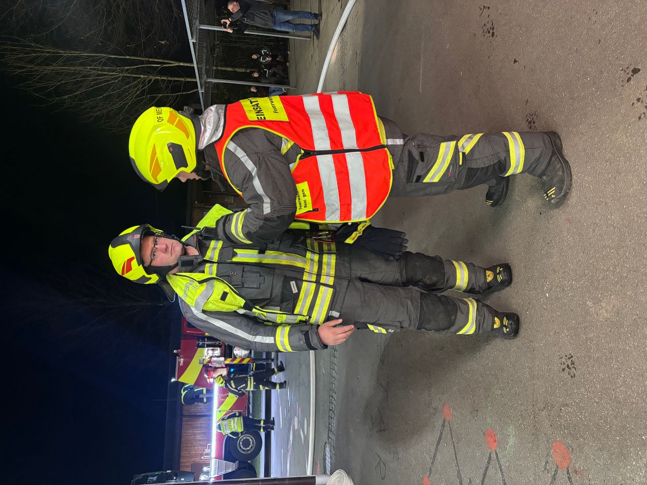 Zwei Feuerwehrleute in reflektierender Kleidung stehen bei Nacht auf einer nassen Straße. Einer trägt einen Helm und Handschuhe, der andere eine reflektierende Weste. Sie stehen neben einem Feuerwehrauto, hinter einer Absperrung befinden sich Zuschauer.